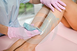 Woman with long tanned perfect legs and smooth skin having wax stripe depilation hair removal procedure on legs in beauty salon.