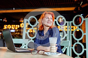 A woman looking away with a smile and a pensive look at a table with a laptop in a cafe