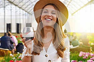 Woman looking at camera and holding glass of wine with people among flowers field drinking and chatting on the background