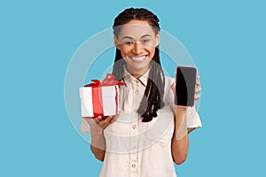Woman looking at camera with toothy smile, showing present box and smart phone with blank screen.
