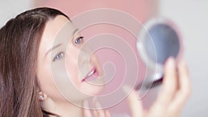 Woman looking at compact mirror and examines her skin, touching face and smiling, long hairstyle and natural makeup