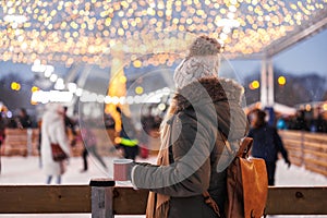 Woman is looking at crowd of people skating on ice rink and drinks hot drink during snowfall