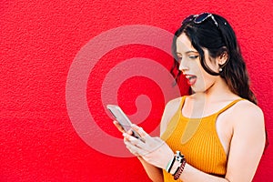Woman looking at her phone with mouth wide open in red background