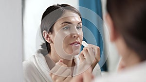 Woman looking at herself in the mirror applies hygienic lipstick or lip balm. In the morning, in a white coat, the girl