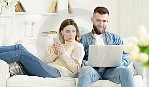 Woman looking at husband`s laptop, sitting together on sofa