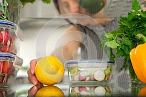 Woman looking inside a fridge full of food and choosing a lemon.