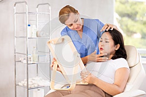 Woman looking at mirror while professional cosmetician explaining her future hardware facial procedure in medical