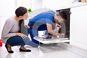 Woman Looking At Repairman Repairing Dishwasher In Kitchen