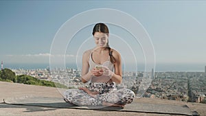 Woman looking at smartphone screen in city. Lady sitting in lotus pose on mat