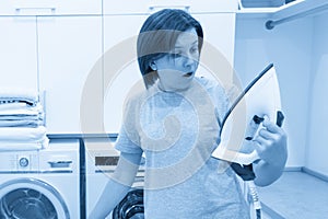 Woman looking with surprise at bottom of broken iron in laundry room with washing machine on background