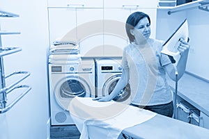 Woman looking with surprise at bottom of iron in laundry room with washing machine on background
