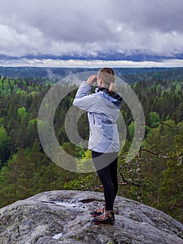 Woman looks through binoculars. tourist girl on a rock in a forest near a lake