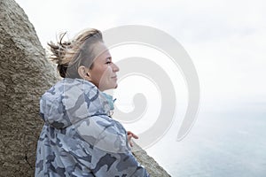 A woman looks down at the sea while standing on a mountain on a sunny day. Cute blonde in a gray jacket. Active recreation in