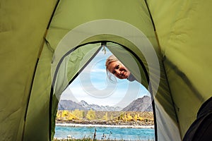 Woman looks inside the green tent and smiles. View from inside tent on the river and mountains on sunny autumn day