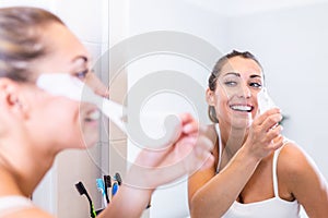 Woman looks in mirror and taking off cosmetic mask. Facial cleansing at home concept. Skin care girl touch patches of fabric mask