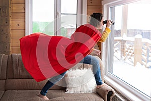 A woman looks out the window with binoculars in a red Cape