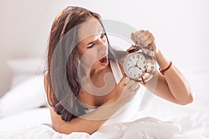 Woman looks shocked at the alarm clock sitting in her white bedding after sleeping in