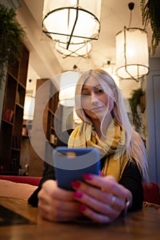 Woman looks at a smartphone in a cafe in the light of artificial cafe lamps