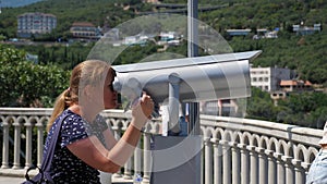 A woman looks through stationary binoculars on an observation deck by the sea.