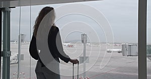 Woman with luggage goes to the airport window to see the plane.