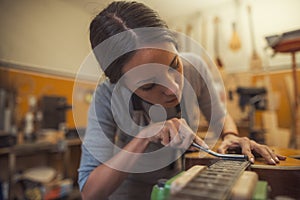 Woman luthier is using a tool to grind a classic guitar fretboard in her musical instrument workshop