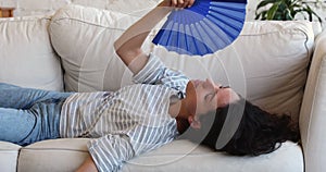 Woman lying on couch feels overheated, wave fan, cools herself