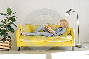 Woman lying on cozy yellow couch and reading book