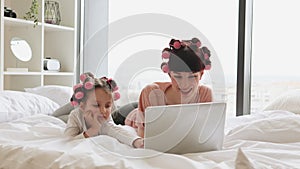 Woman lying with a cute girl on the bed, curling their hair, using a laptop.