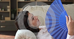 Woman lying on sofa holds fan, cools herself, feels exhausted