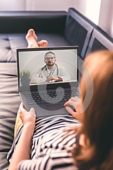 Woman lying on a sofa and talking with a doctor online using laptop. Telemedicine concept.