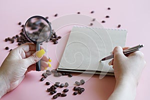 Woman with magnifying glass, notebook and pen writes down the results of the analysis of the quality of roasted coffee beans in a