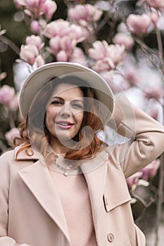 Woman magnolia flowers, surrounded by blossoming trees., hair down, white hat, wearing a light coat. Captured during