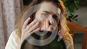 Woman without make up applying cosmetic patches under eyes looking in mirror.