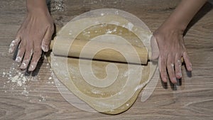 A woman makes pizza at home. She flattens a piece of yellow dough on the wooden table with a rolling pin. The concept of