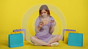 A woman makes a purchase in an online store using a smartphone and a credit card. Redhaired woman sits crosslegged on