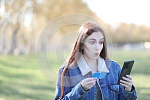 Woman makes a surprised gesture while holding her credit card and looking at her cell phone making a purchase