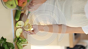 Woman making detox beverage at home. Organic products on wooden table. Female slices a lemon. Apple, kiwi, celery, and
