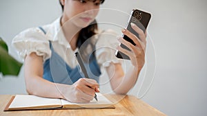 A woman making lists in her notebook while using her smartphone, sitting at a table indoors