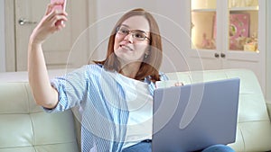 Woman making selfie sitting on couch and verifies social network on laptop.