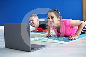 Woman and man do push-up on gymnastic mat and watch video in loptop.