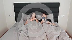 Woman and man lying in bed with smartphones in hands. Young married couple on grey bed watching at devices and texting in networks