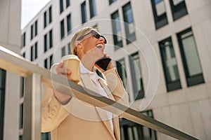 Woman manager talking phone whiile standing on modern building background and drinking coffee