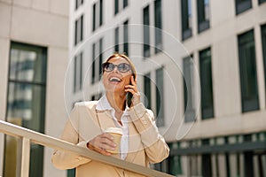 Woman manager talking phone whiile standing on modern building background and drinking coffee