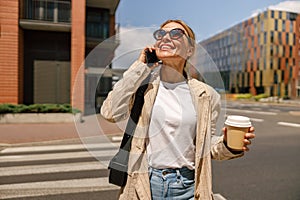 Woman manager talking phone whiile standing on modern building background and drinking coffee