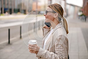 Woman manager talking phone whiile standing on modern building background and drinking coffee