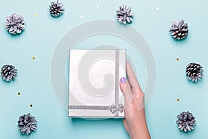 Woman manicured hands holding silver gift box on blue background with pine cones and confetti, copy space, top view, flat lay.