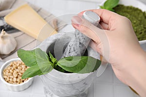 Woman mashing basil for pesto sauce in mortar at white table, closeup