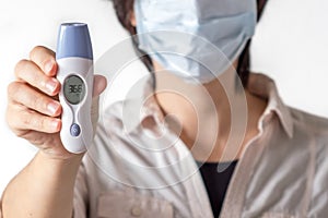 A woman in a mask holds an electric clinical thermometer. The thermometer shows that the woman is healthy