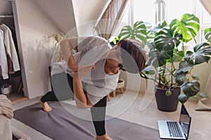 A woman on a mat doing twist and stretching asana position in her bedroom. Online class yoga practice at home. Healthy