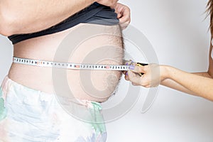 A woman measures an overweight man& x27;s belly with a measuring tape on a light background, obesity and diet